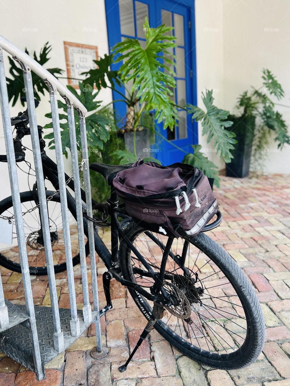Road bicycle with weathered pack chained to a spiral staircase, near a building entrance. It looks like a working bike, waiting to deliver a package.