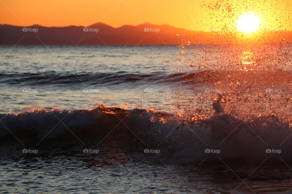 Ocean , mountain and setting sun and wave crashing and exploding in tiny golden drops