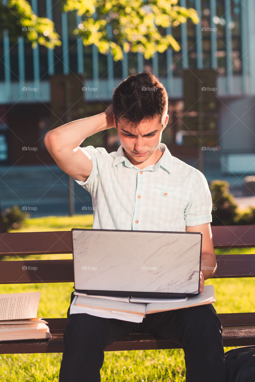 Student working on a laptop using books and notes sitting on a bench in a park. Young boy wearing a blue shirt and dark jeans