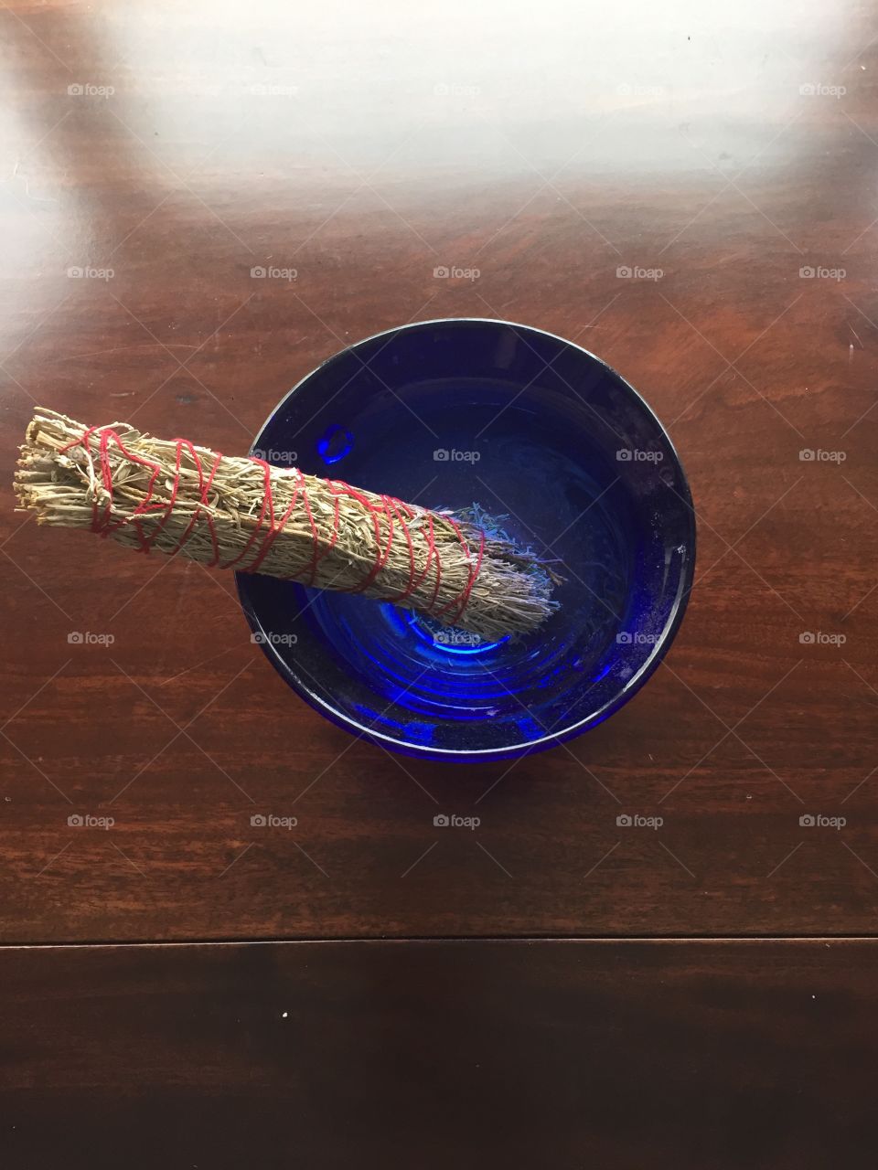 A large sage smudge sits in a blue glass bowl on a dark wood table. Morning sunlight illuminates the picture from the top.