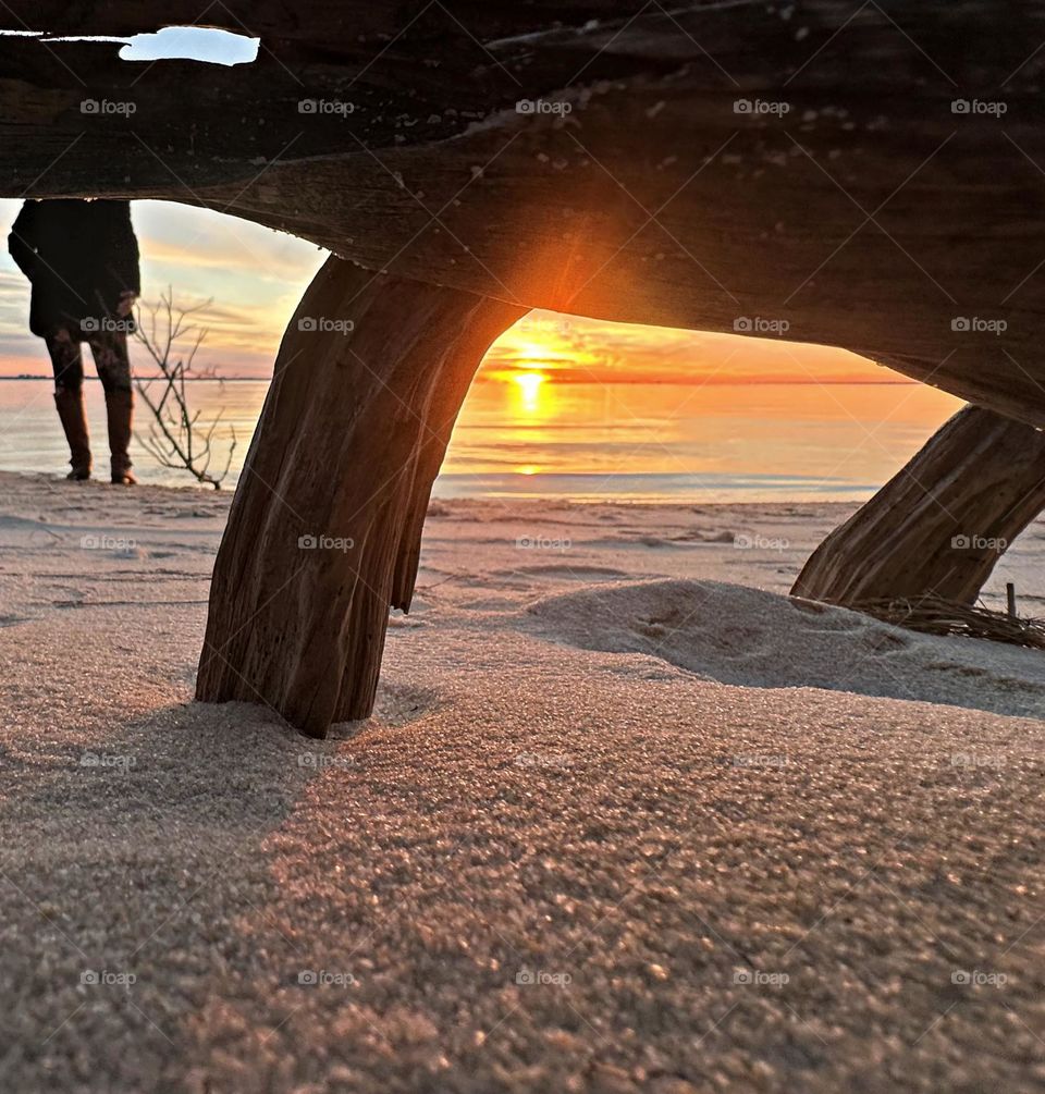 A descending sunset reflection off the waters surface. Taken from underneath a buried log in the sugary white crystal sand as a young women takes photos. 