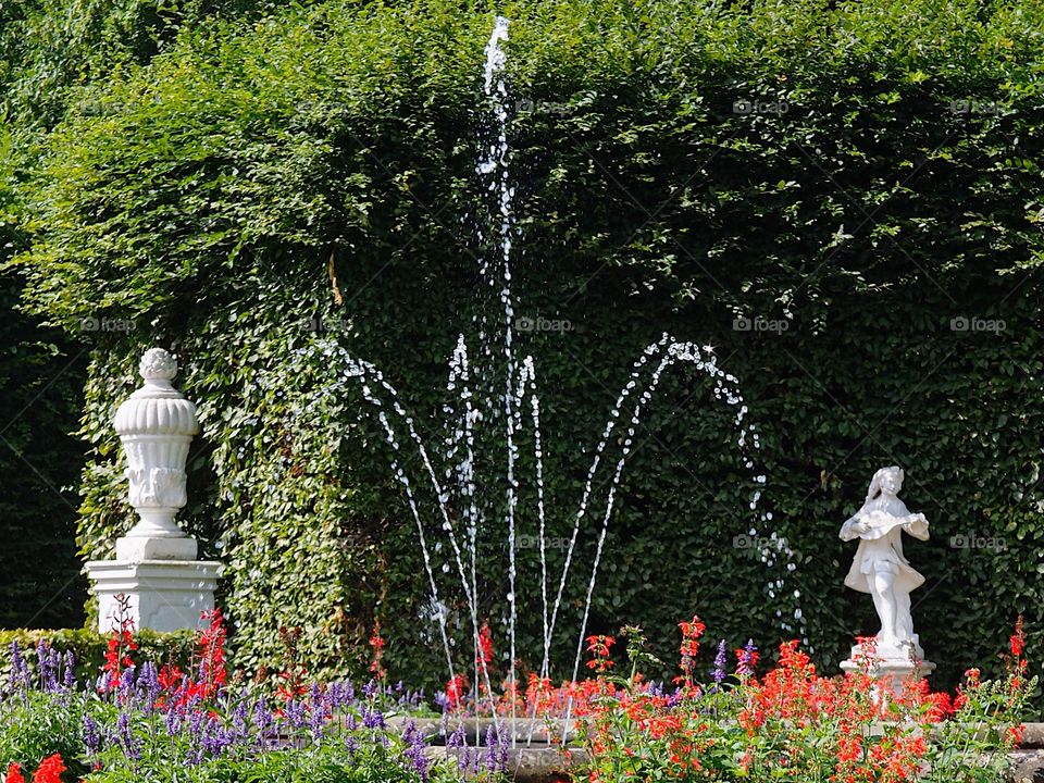 A beautiful outdoor fountain surrounded by bright red, pink, white, and purple flowerbeds along with statues and finely manicured hedges in the background in a public park in Europe on a sunny summer day.