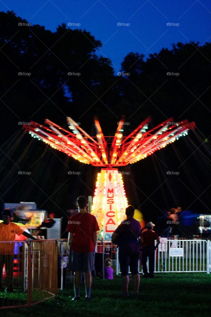Multicolored lights of a ride at a fairground at night with silhouettes of people watching