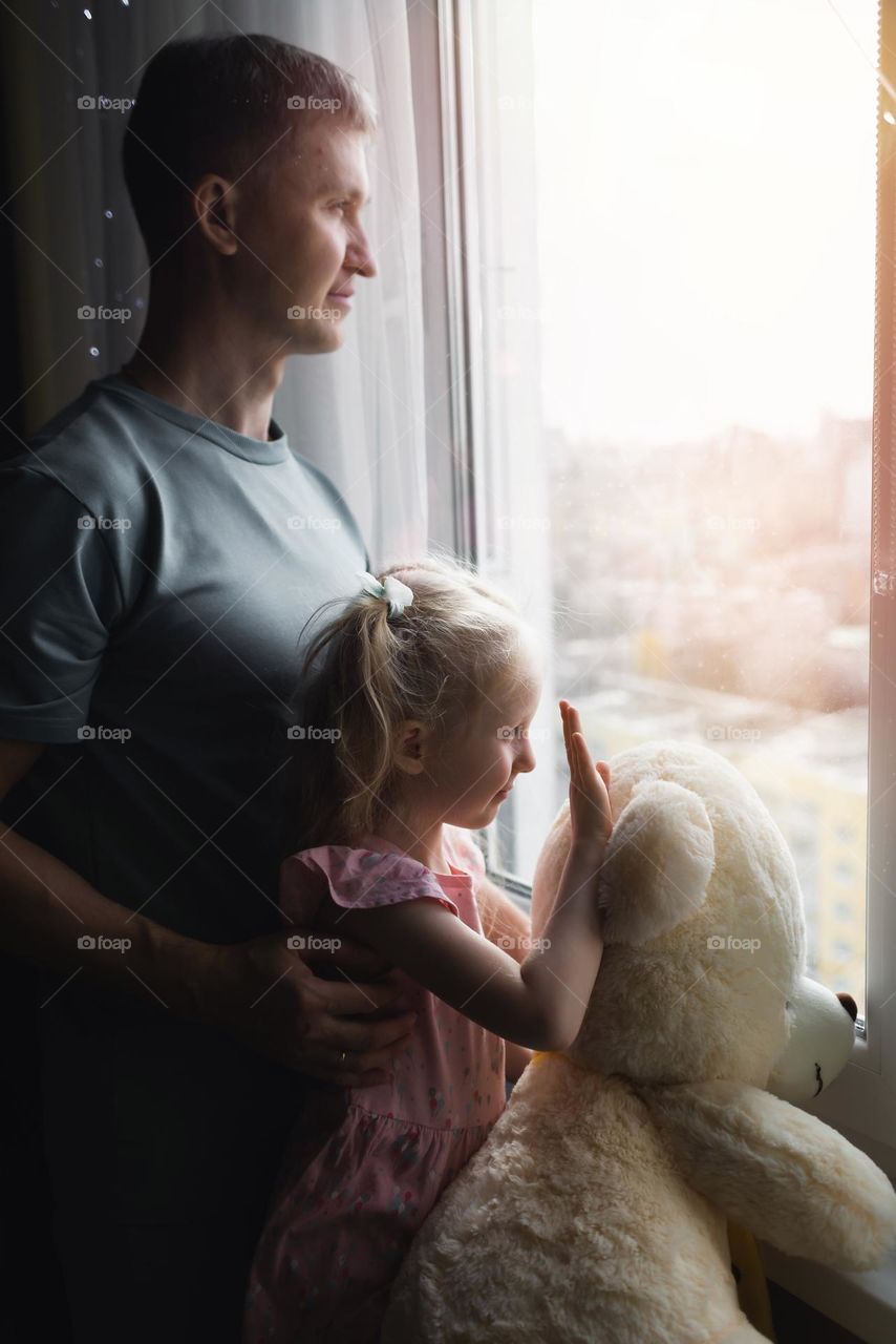 Side view portrait child girl and dad look out the window at home