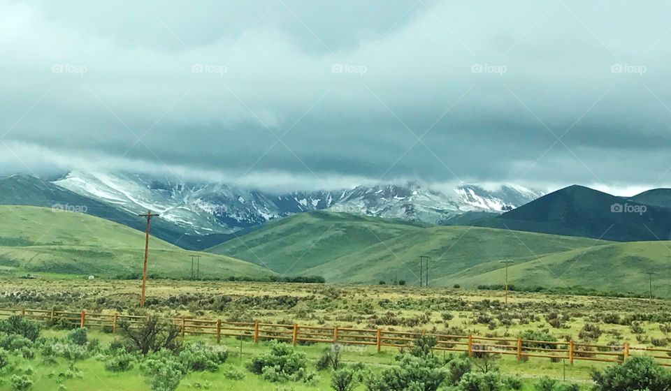 Lovely summer country landscape with green shrubs, grass, a wooden fence, hills, snowy mountains, and a grey stormy sky. 