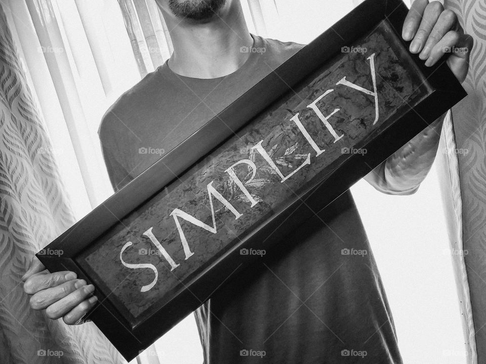 In nlack and white, a man holding a framed encouraging word art piece with the word simplify.