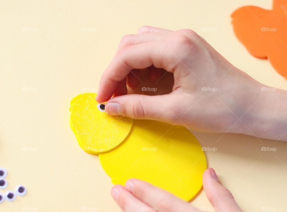 The hands of a little Caucasian girl glues her eyes on a yellow felt chicken,sitting at a table in her room, close-up side view. Creative kids concept, easter preparation.