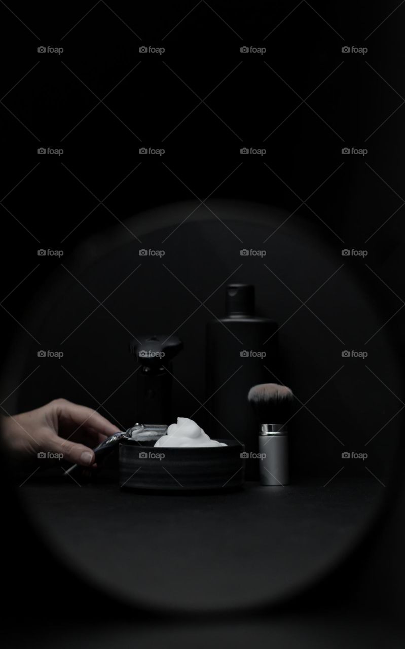 The hand of a caucasian young woman holds a razor over a bowl of shaving foam, and next to it a shaving brush and bottles of cosmetics are reflected in a round small mirror on a black background, close-up side view