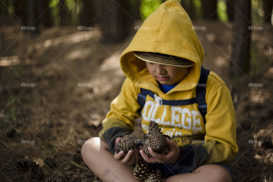 Seasonal outdoor portrait of a young happy Eurasian kid hiking in a pine forest wood.Natural setting, the boy is wearing yellow sweater with hood and cap on. Alone, seating on the floor holding pinecones with both hands