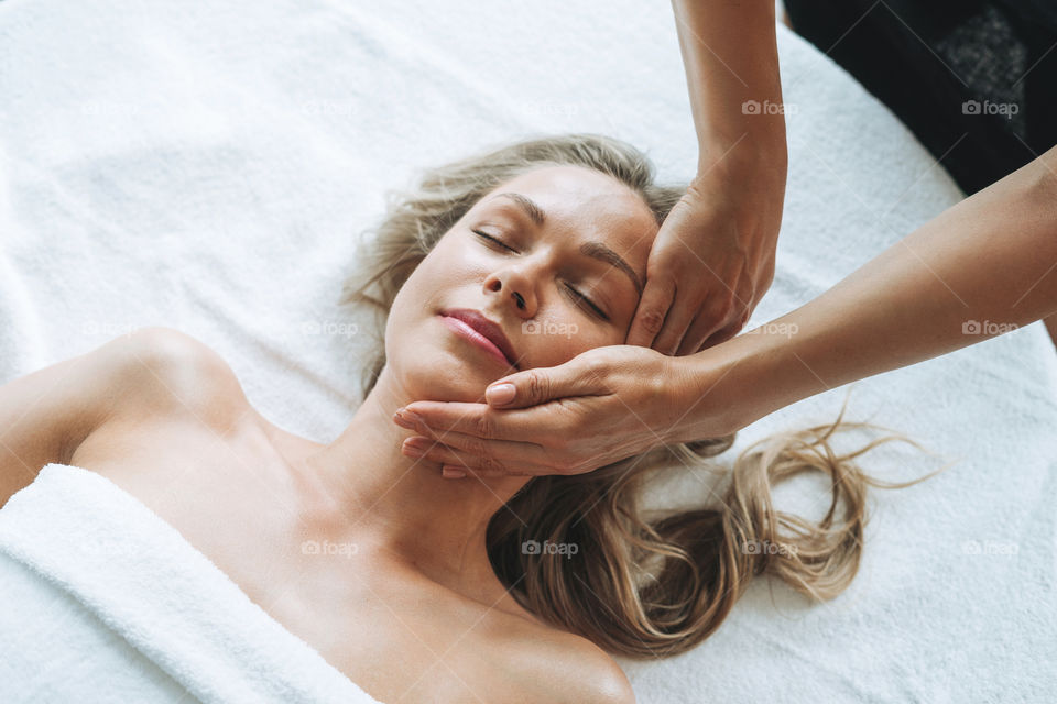 Young blonde woman gets facial massage by hands in beauty salon spa
