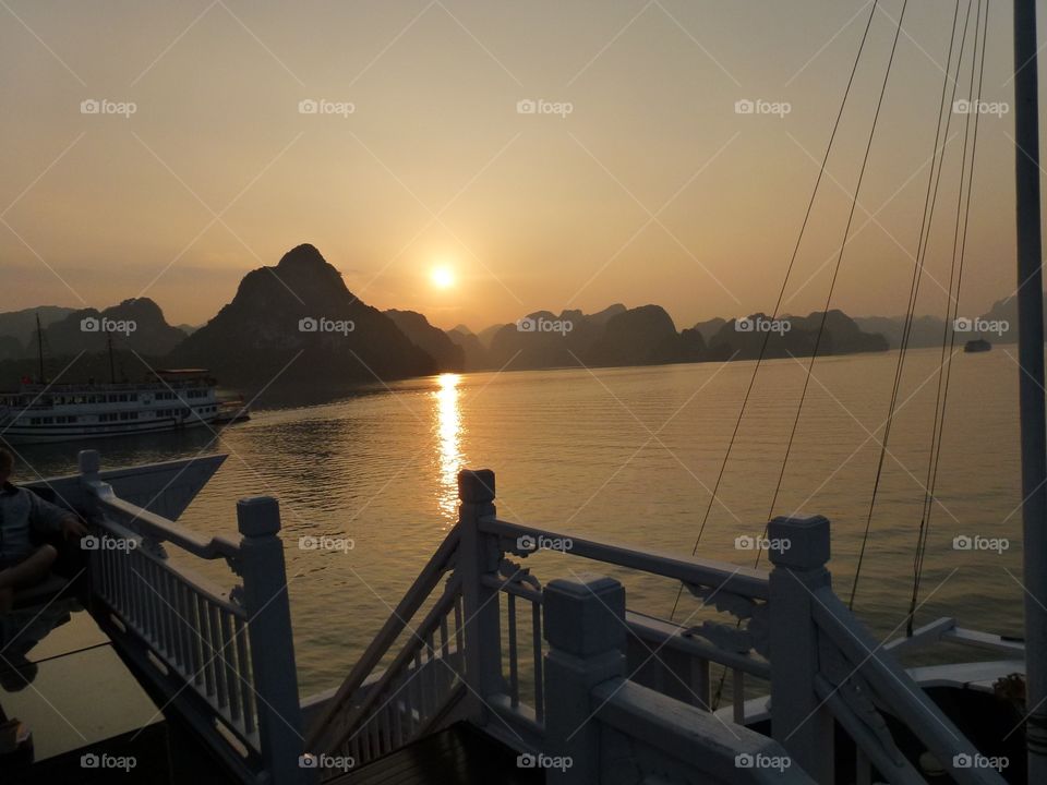 the surrounding darkened, the black mountains , the white wooden slats on the yacht also turned black, accentuating the orange sun in the sky. the colors reflect on the water, the scene is so romantic