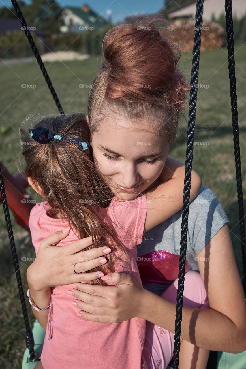 Teenage girl hugging her younger sister in a home playground in a backyard. Happy smiling sisters having fun on a swing together on summer day. Real people, authentic situations