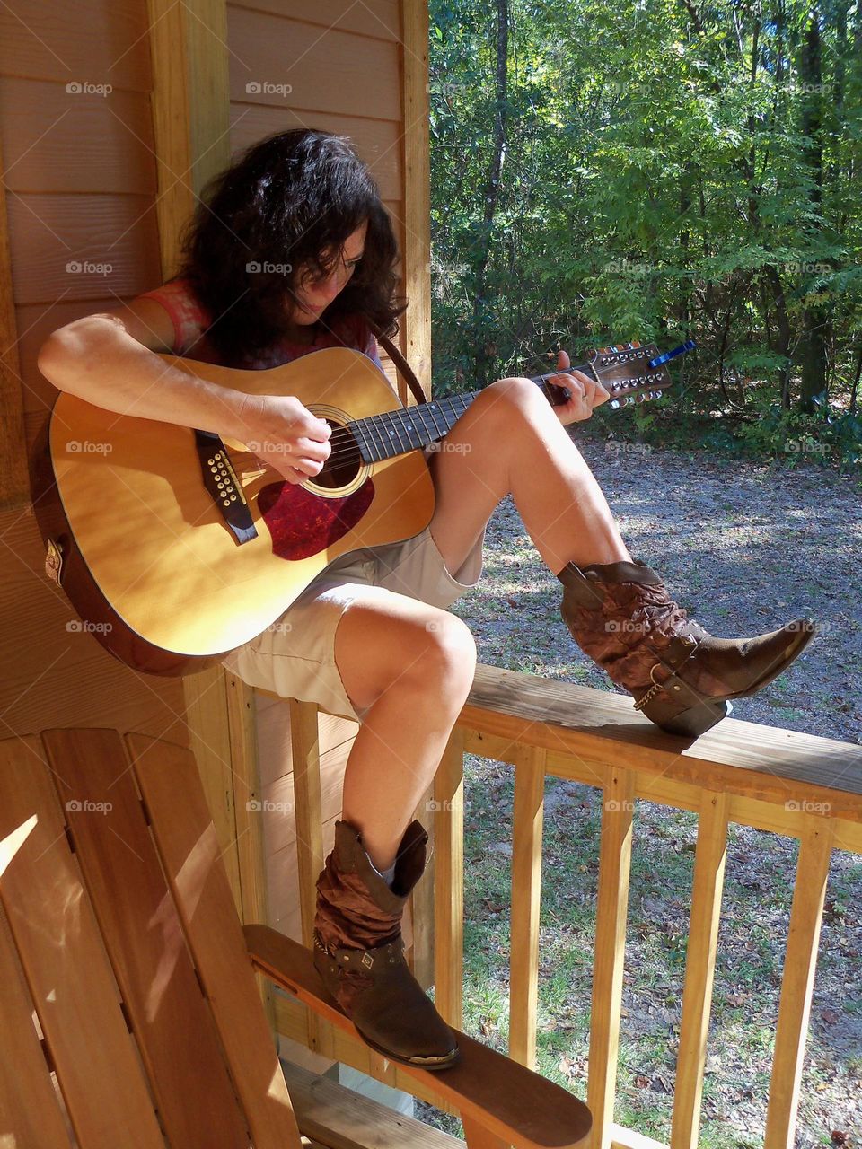 Woman sitting on balcony wooden rail wearing cowboy boots playing a twelve string guitar