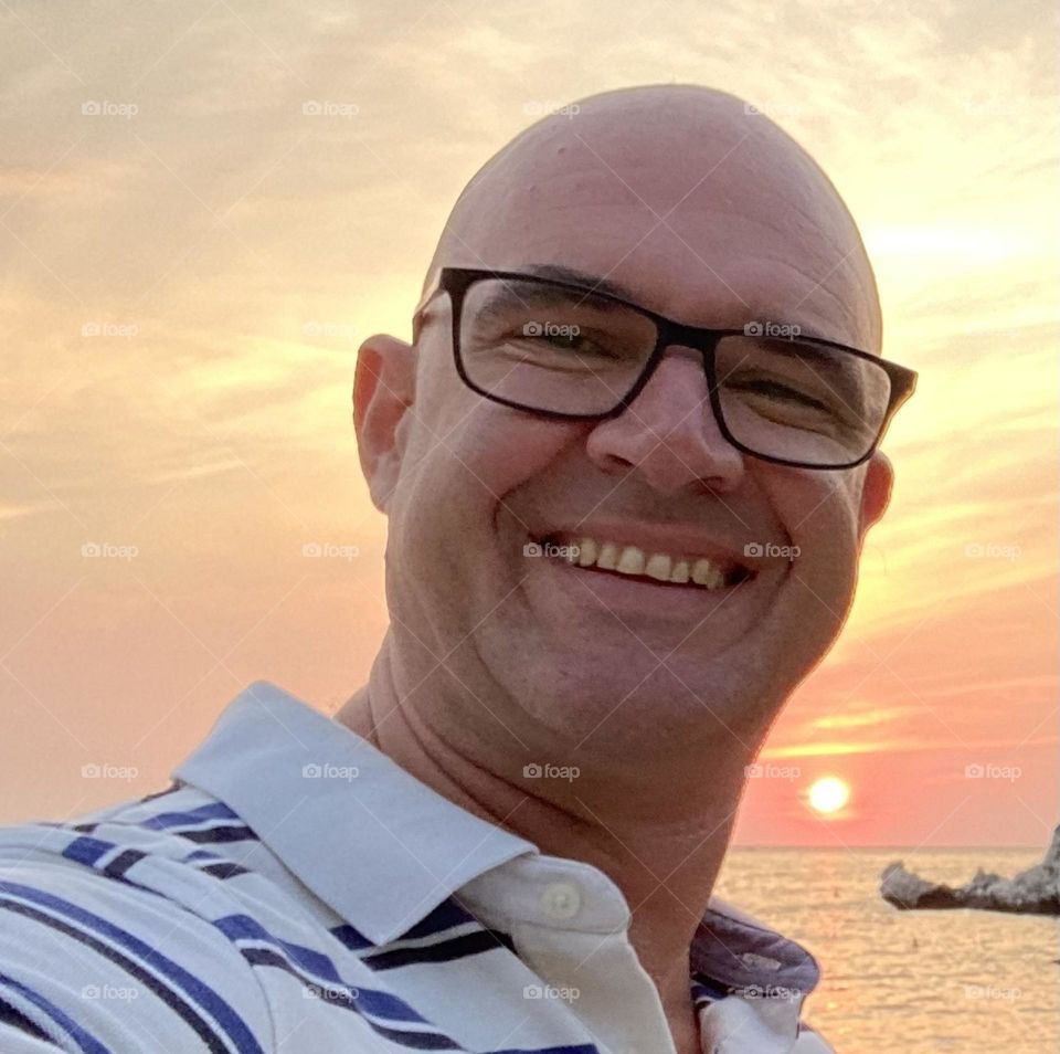 A smiling bald headed man with glasses with the background of the setting sun and the Adriatic Sea.