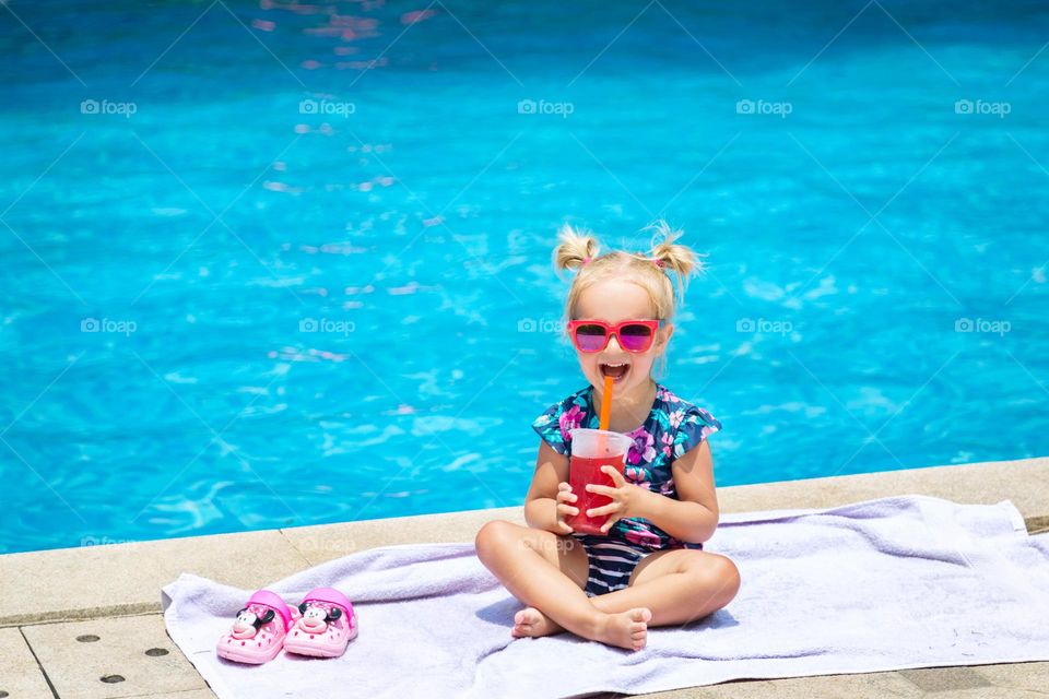 Happy little Caucasian girl drinking juice in swimming pool 