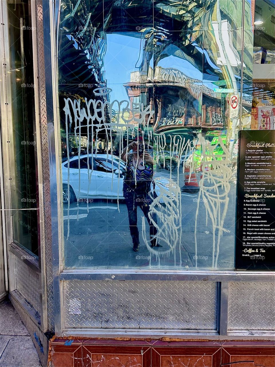 The busy intersection of “Jefferson St”, “B’way” & “Myrtle Ave” in “Bushwick”, Bklyn by the “M” train overpass and station is visible in a slightly warped white graffiti covered mirror as well as myself taking the picture. 2024. Hypnotic Productions