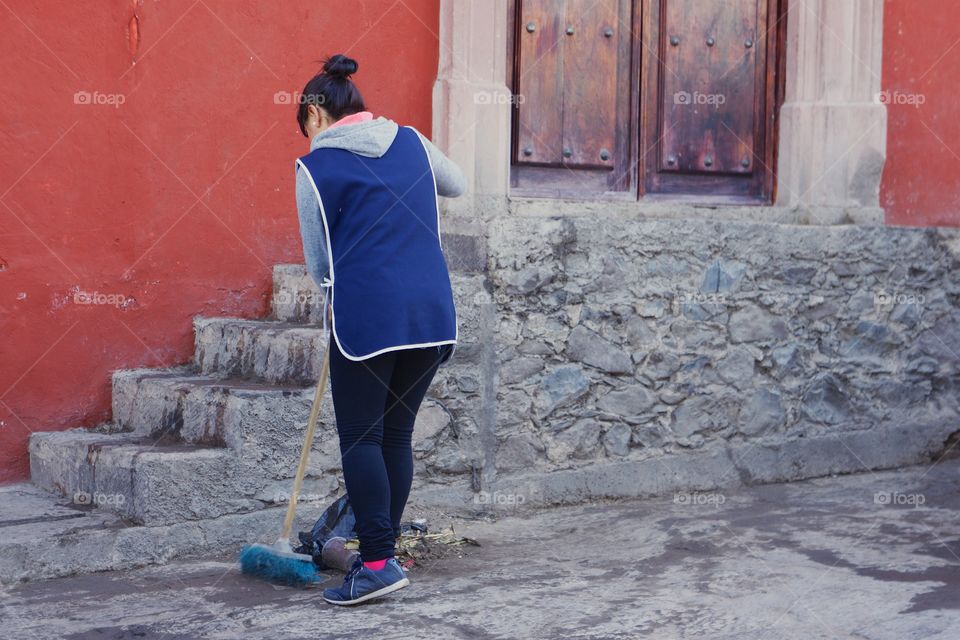 A woman sweeping rubbish and dirt outside the exterior of a home where she works as a maid

