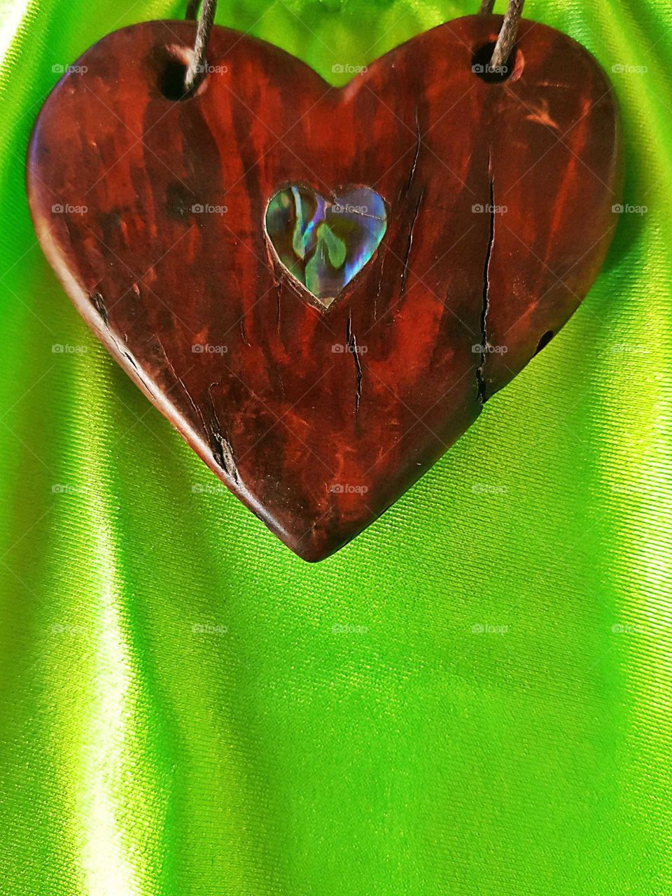 A piece of driftwood shaped like a heart. Polished with a piece of Paula shell made into a heart shape inserted into the centre. A unique necklace. Sitting on a bright green background.