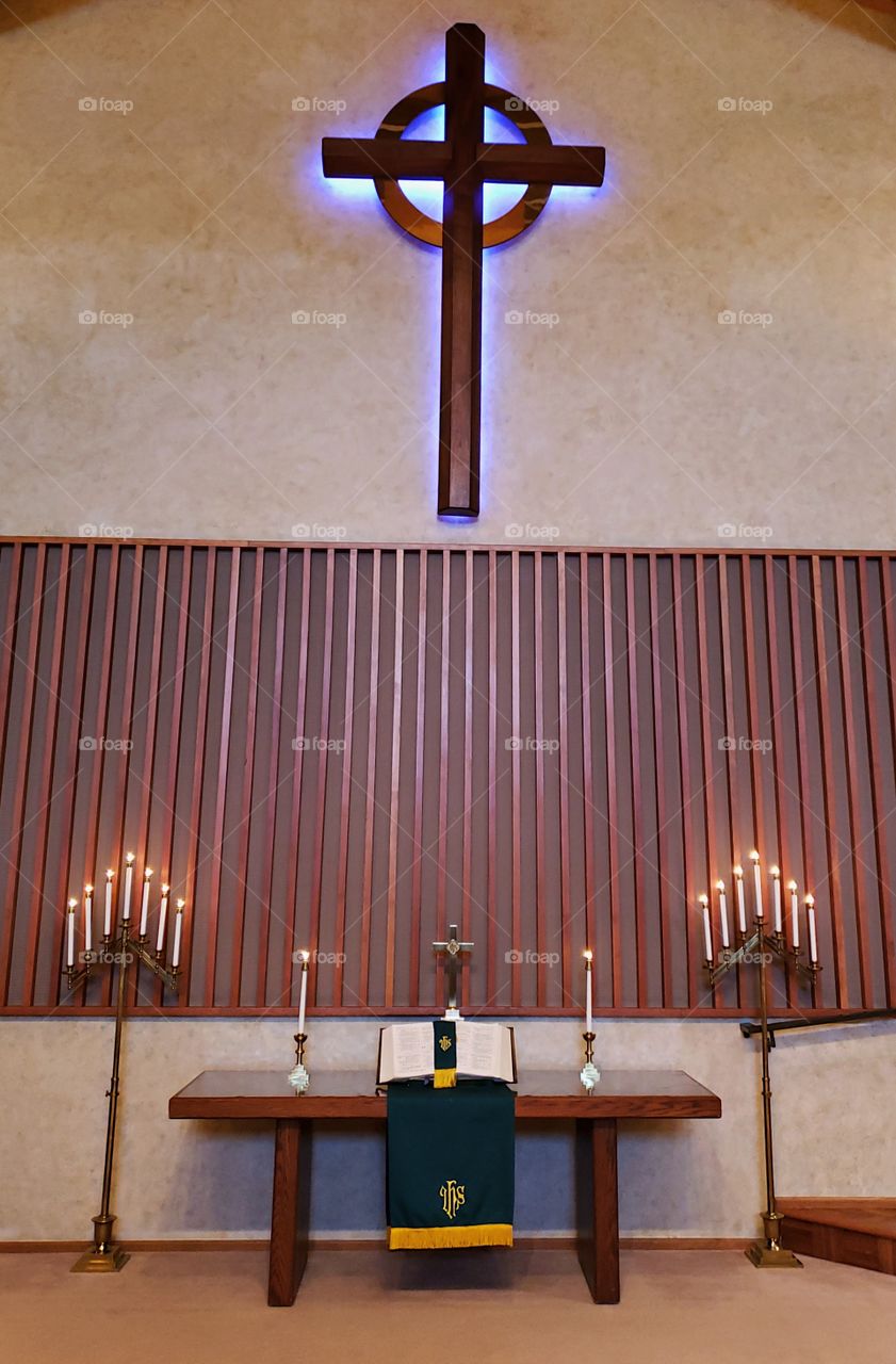 An alter setting with a beautiful table with candles on the side, a large open bible on a stand, and green and yellow paraments draped from the table with a large wooden cross above with a purple glow. 
