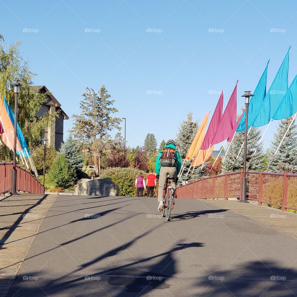 A couple walks for morning exercise while a bicyclist rides on his morning commute across a bridge adorned with colorful flags at the Old Mill District in Bend in Central Oregon on a sunny fall day.