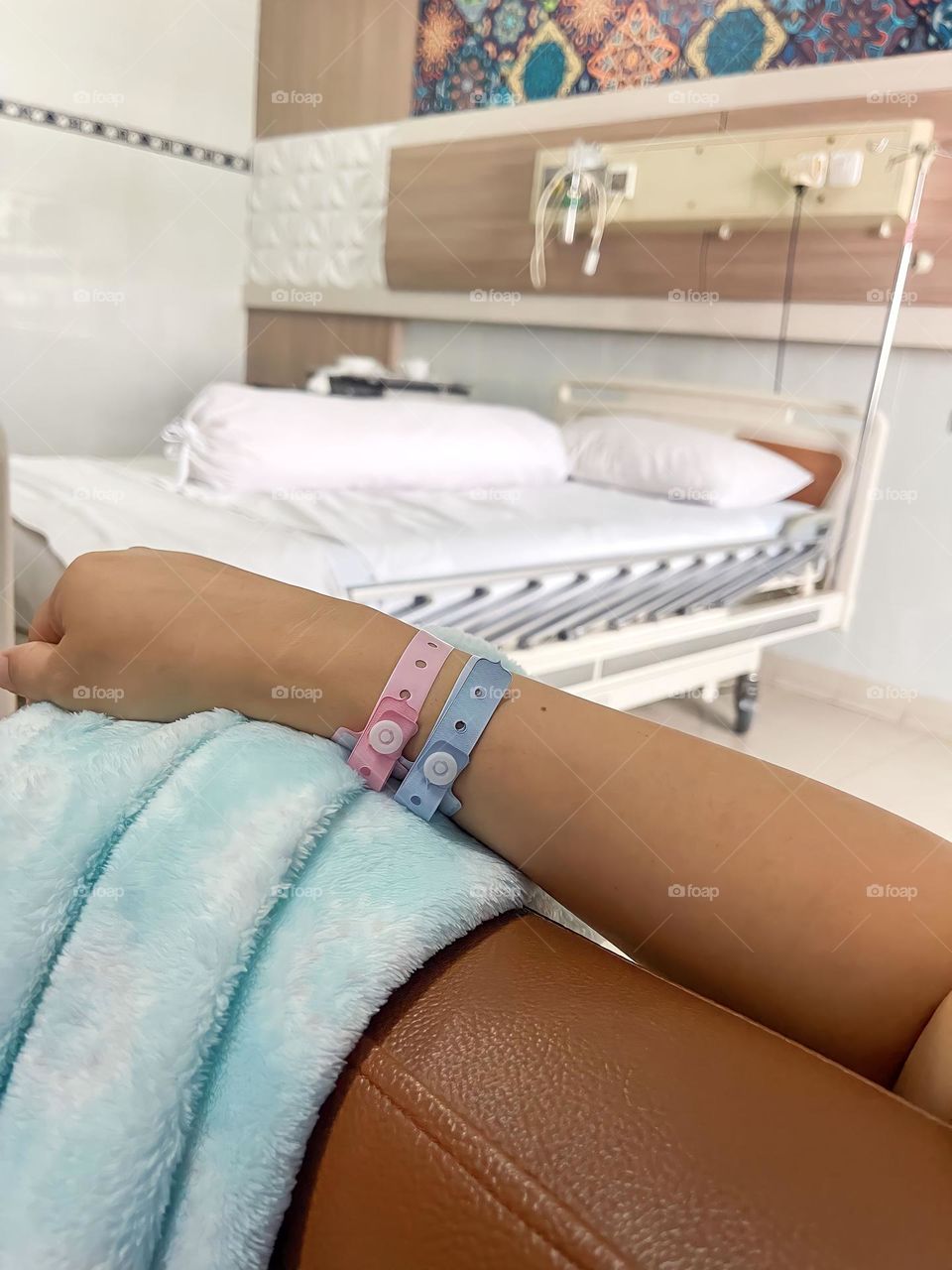 A baby's hand with a pink identification bracelet lies on a light blue blanket in a hospital room