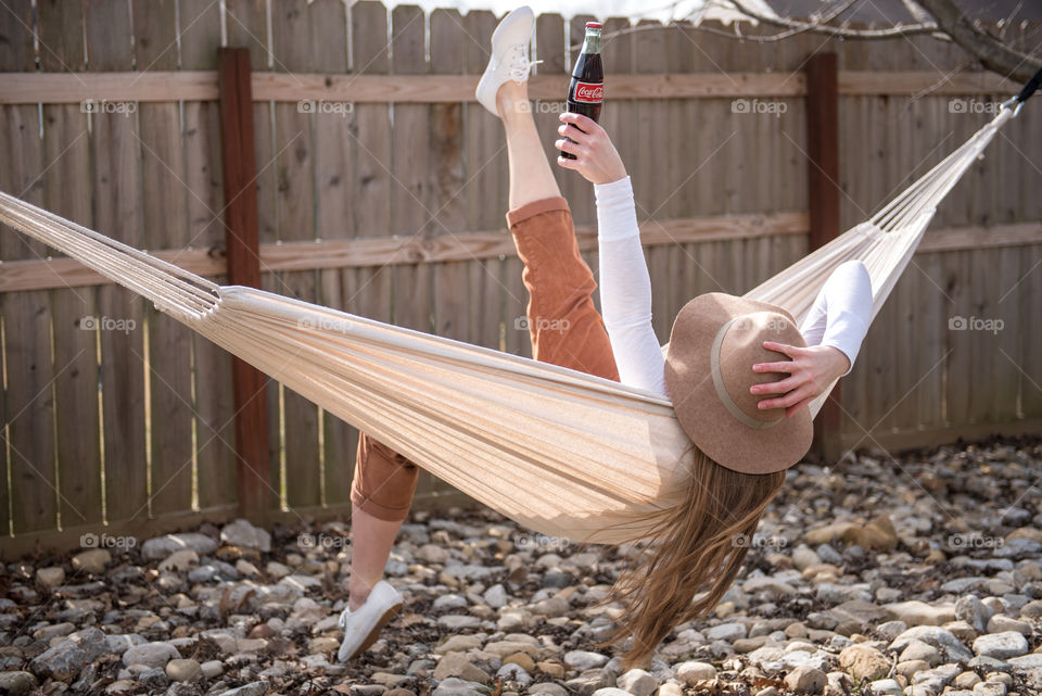 Young millennial woman swinging in a hammock and holding a bottle of Coca-cola in the air