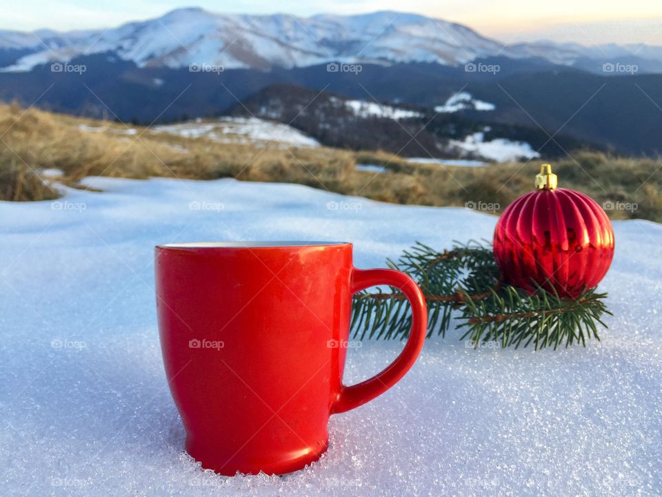 Red mug placed on snow with red globes and evegreen branches beside and snowy mountains in the background