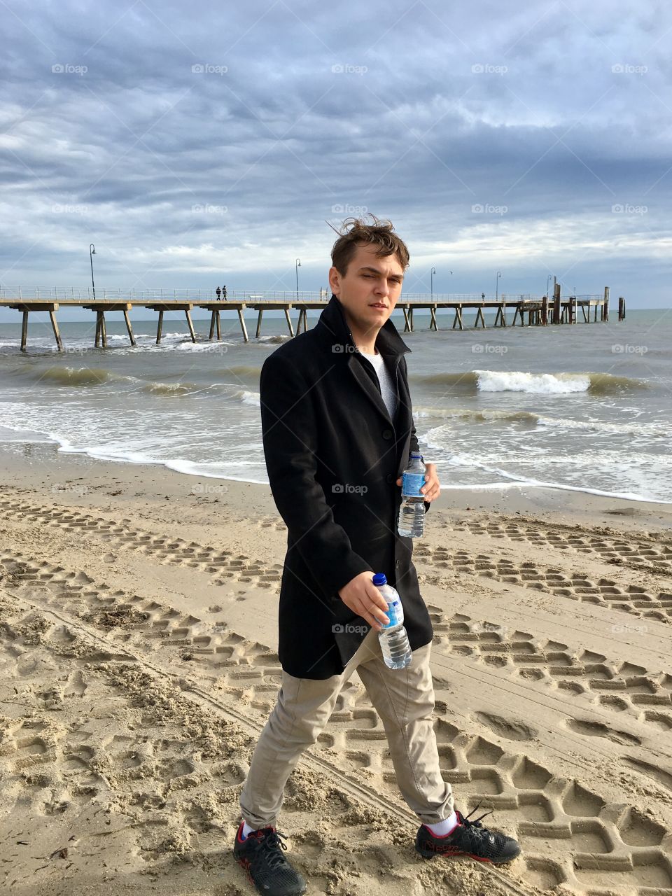 Handsome young man wearing black wool coat and holding two bottles of water walking on beach 