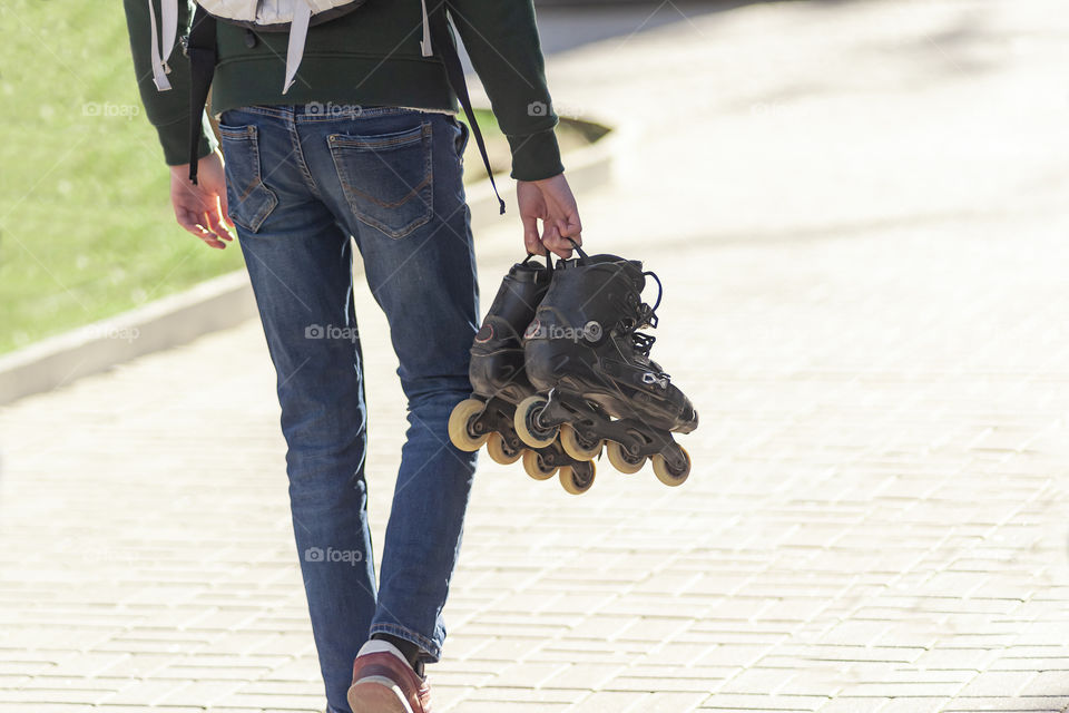 young guy walks down the street on a sunny spring day and carries roller skates in his hand