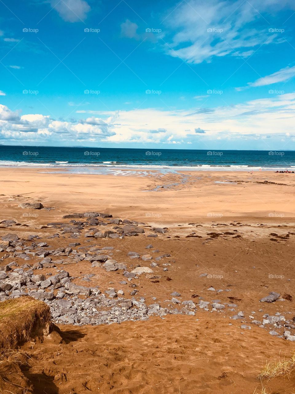 Fanore Beach in County Clare, Ireland, offers a striking mix of dark rocky sand, smooth pebbles, and light granular sand. The beach curves toward the crashing Atlantic waves under a bright blue sky, creating a serene yet untamed coastal scene.
