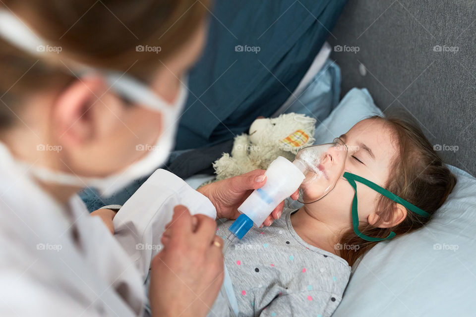 Doctor visiting little patient at home. Child having medical inhalation treatment with nebuliser. Girl with breathing mask on her face. Woman wearing uniform and face mask