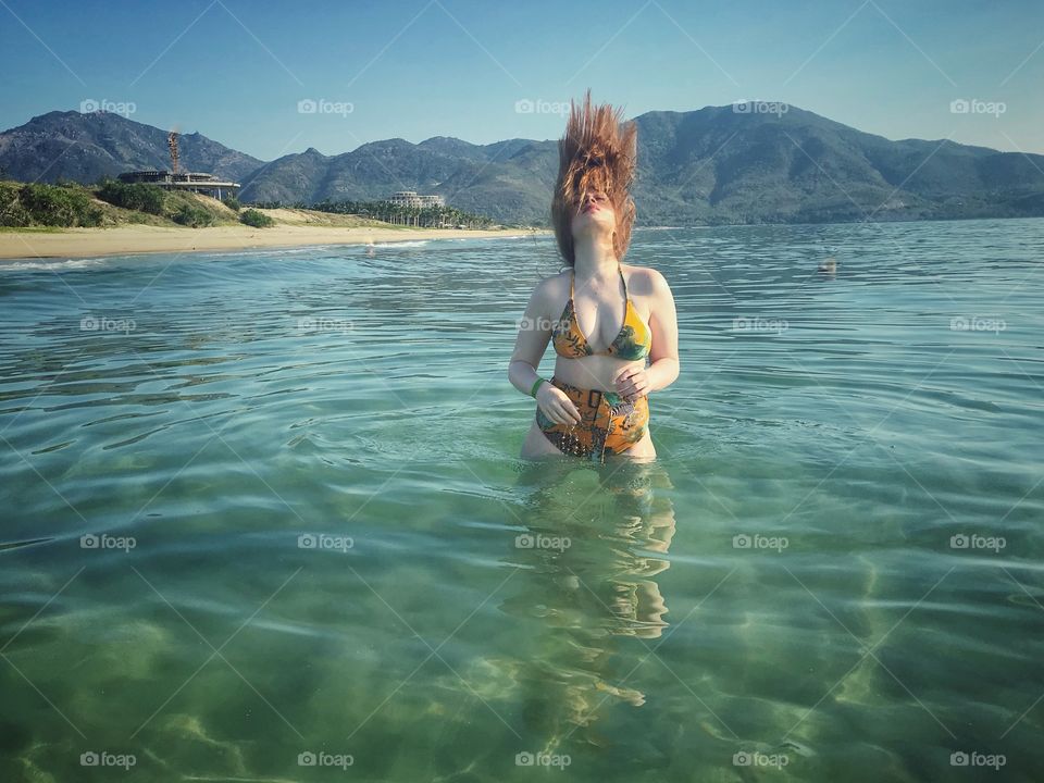 a girl with red hair in a swimsuit is standing in the sea up to the waist, clear sea, clear water, against the background of mountains, throws her hair back, plays with her hair, seascape, water landscape, summer, vacation, Relax.