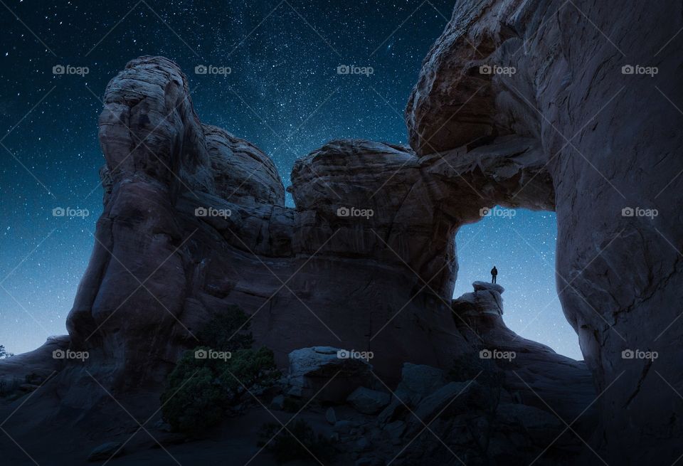 A man looks off at the stars while standing under a massive natural arch in the Utah desert