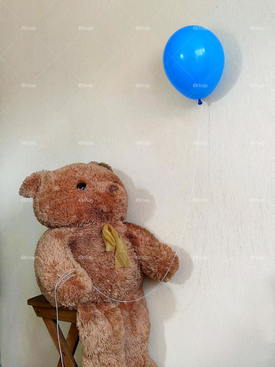 Minimalism of objects in space.  The Teddy bear sits on a wooden chair and looks at the blue balloon he is holding by the rope.  The background is pale beige.