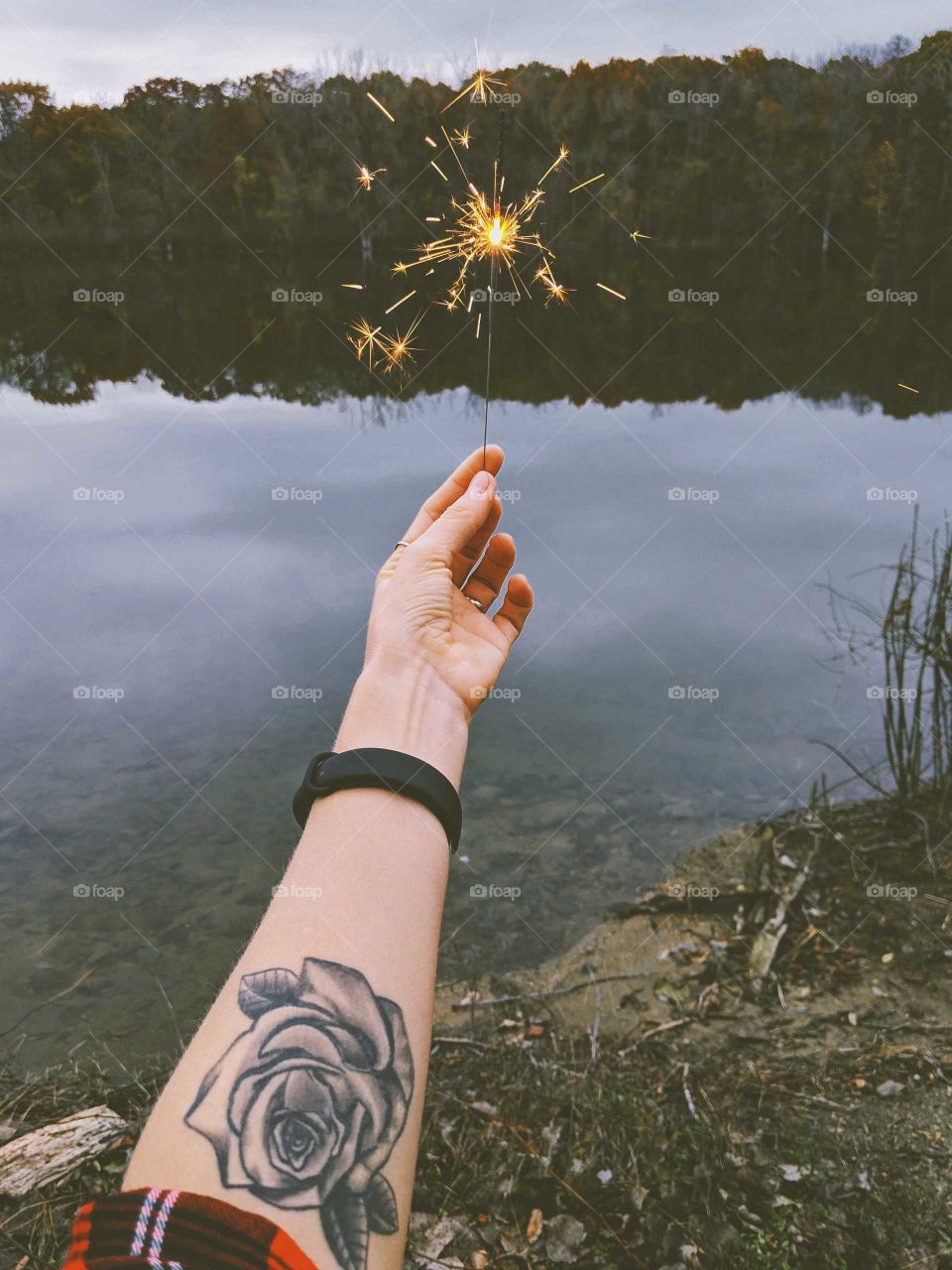 A hand holding up a sparkler, with a view of a lake and an autumn landscape in the background