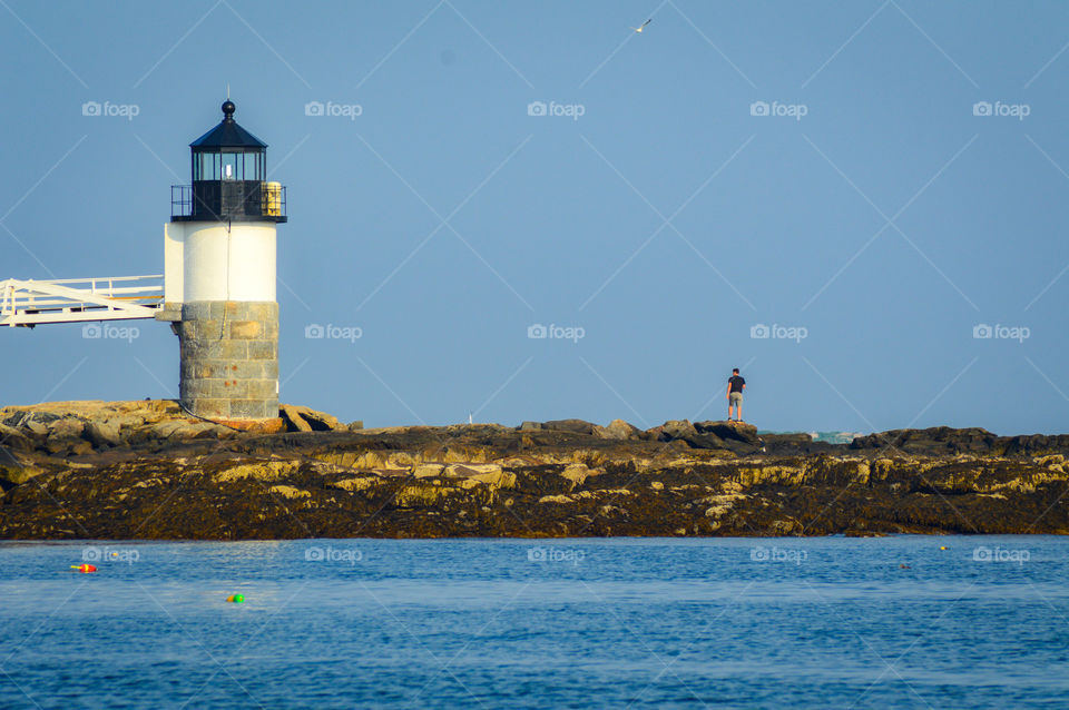 Lighthouse on the Maine coast with a man standing nearby, and water in the foreground.