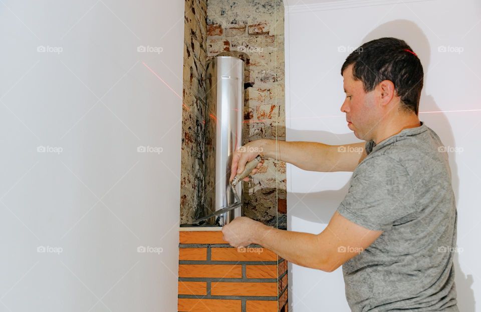 Portrait of a young, handsome Caucasian brunette man applying fresh cement mortar to the brick of a fireplace chimney, holding and trimming with a narrow wooden lath, standing on a chair in the evening, close-up view from the side. Concept clean bric