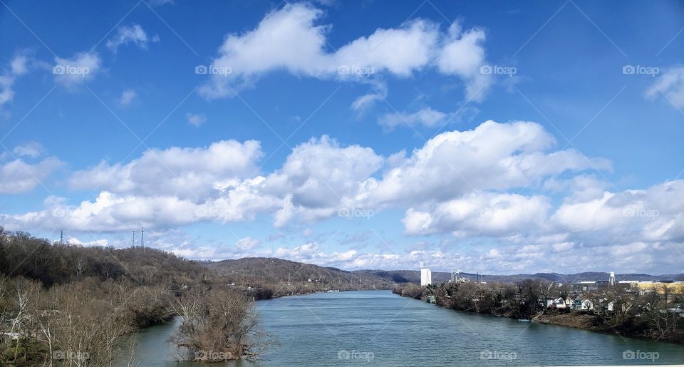 White fluffy clouds in bright blue sky over river with trees and mountains