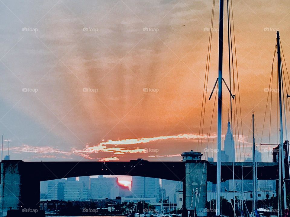 The sun is setting behind the horizon at Newtown Creek in Long Island City, Queens, NY and casts its orange red golden light into the misty slightly overcast sky creating a rippled shape like a crown. 2018. Hypnotic Productions