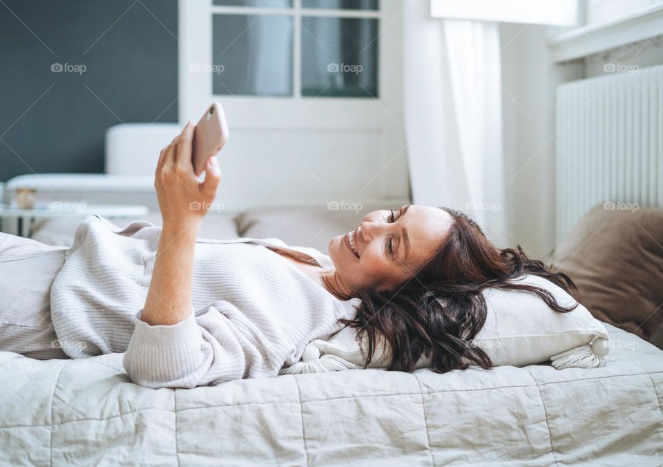 Young woman forty year with brunette long hair in cozy knitted cardigan using mobile phone in bed at home