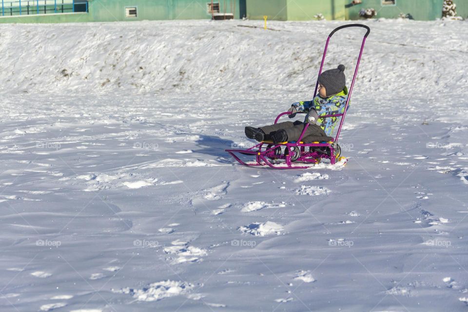 A child in winter in winter jackets, pants, a hat and boots on white snow on the street and in the park in nature plays winter fun and sleds.