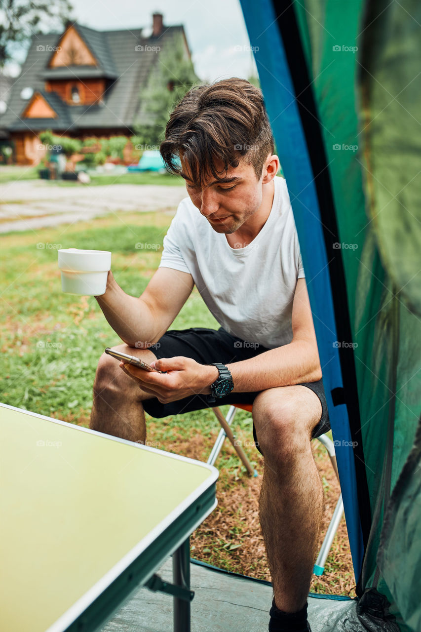 Young man drinking a coffee sitting at front of tent in the morning. Teenager enjoying free time during weekend trip in summer