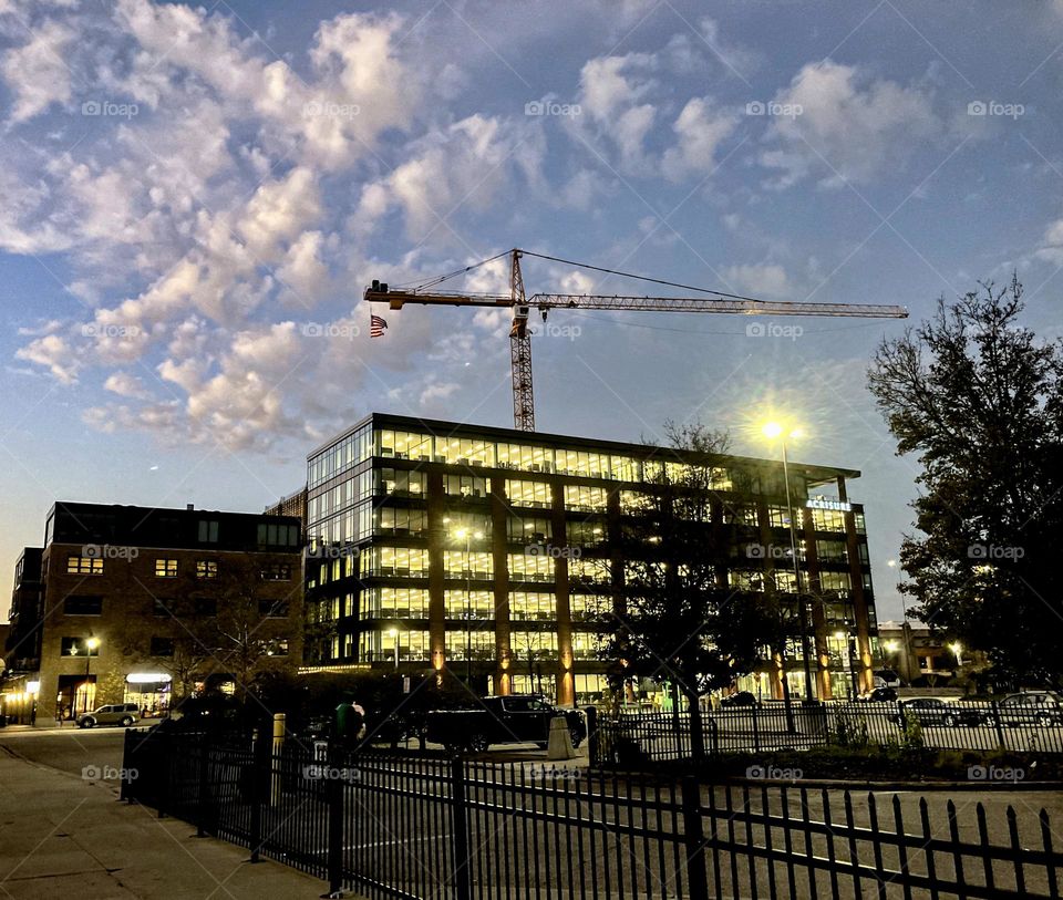 Building under construction has a crane adorned with an American flag above it and is all lit up as the sun just starts to rise lighting up the sky in bright blue with white clouds in the city in downtown Grand Rapids Michigan