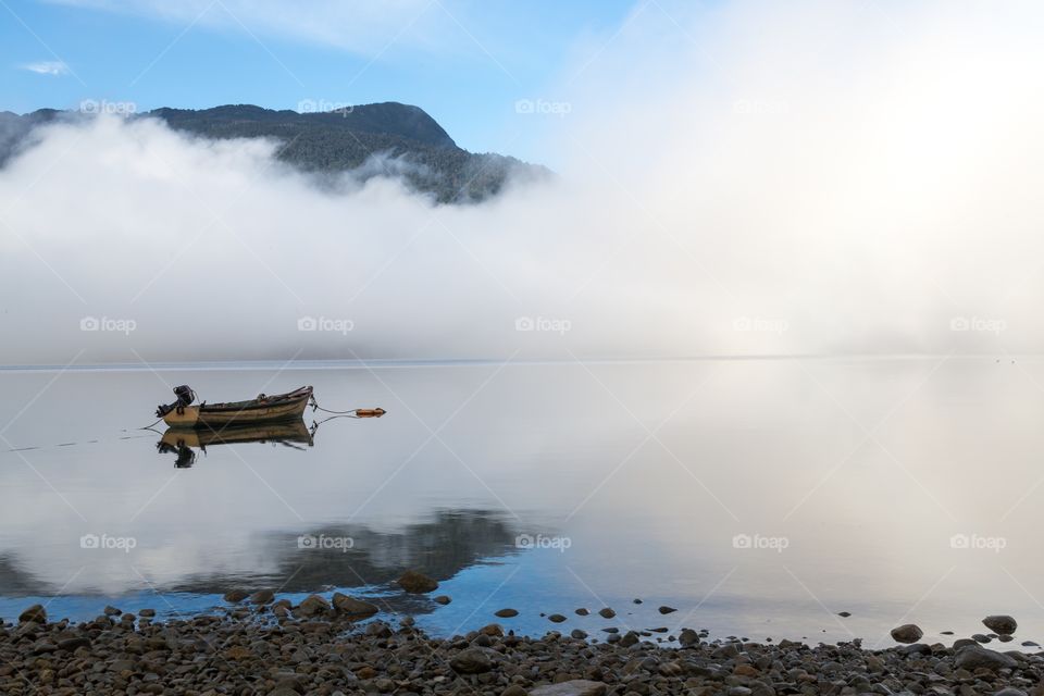 Small boat on a foggy sea. Small rowing boat on a foggy sea during sunrise. Mountain in the background is visible. Shoreline in the front