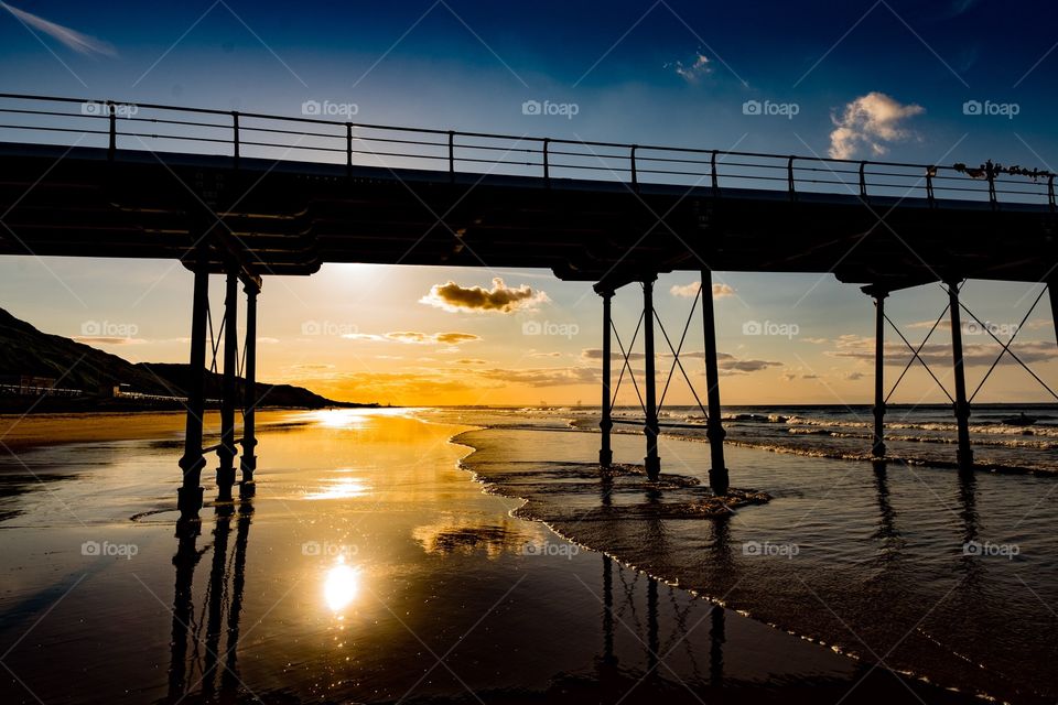 Sunset under the pier. The Victorian pier at Saltburn in the UK is silhouetted against a golden sunset, which is reflected in the sea below. 