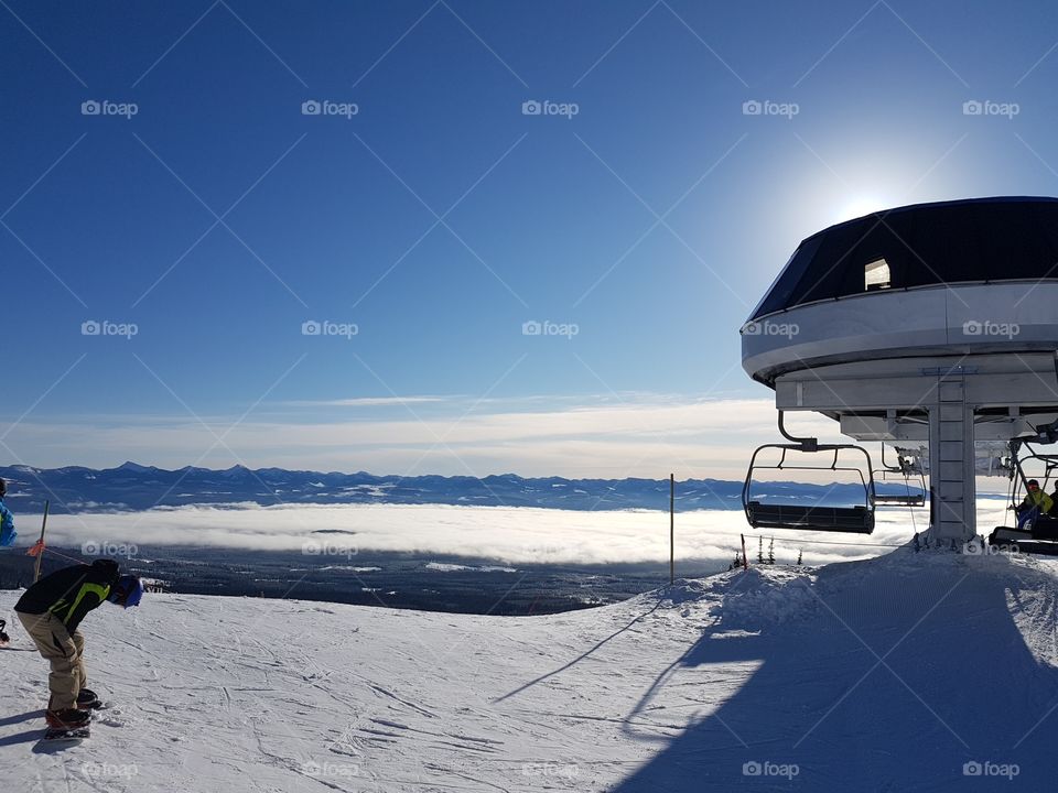 Ski lift at the top of the mountain with inverted cloud layer in the valley in the distance. Blue skies with a few clouds.