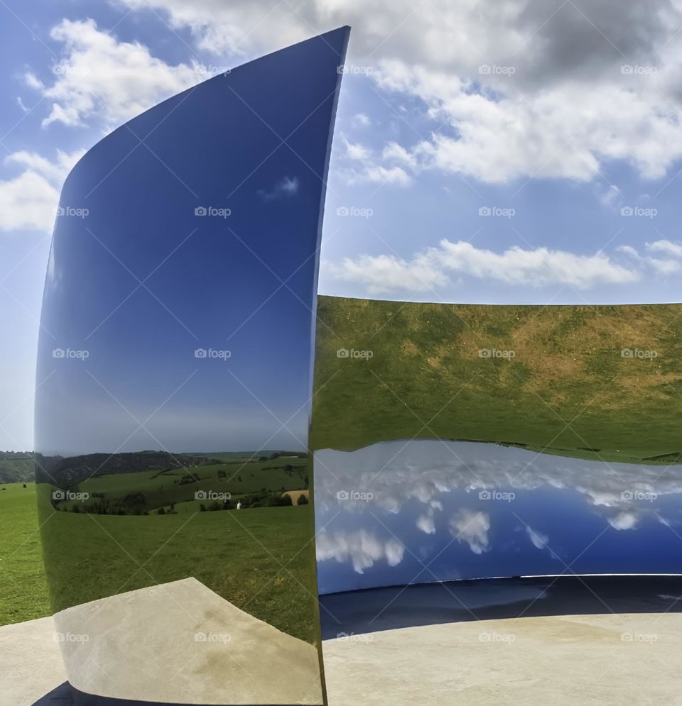 Blue sky with fluffy white clouds and green fields reflected in a curved mirrored structure, turning the scene upside down 