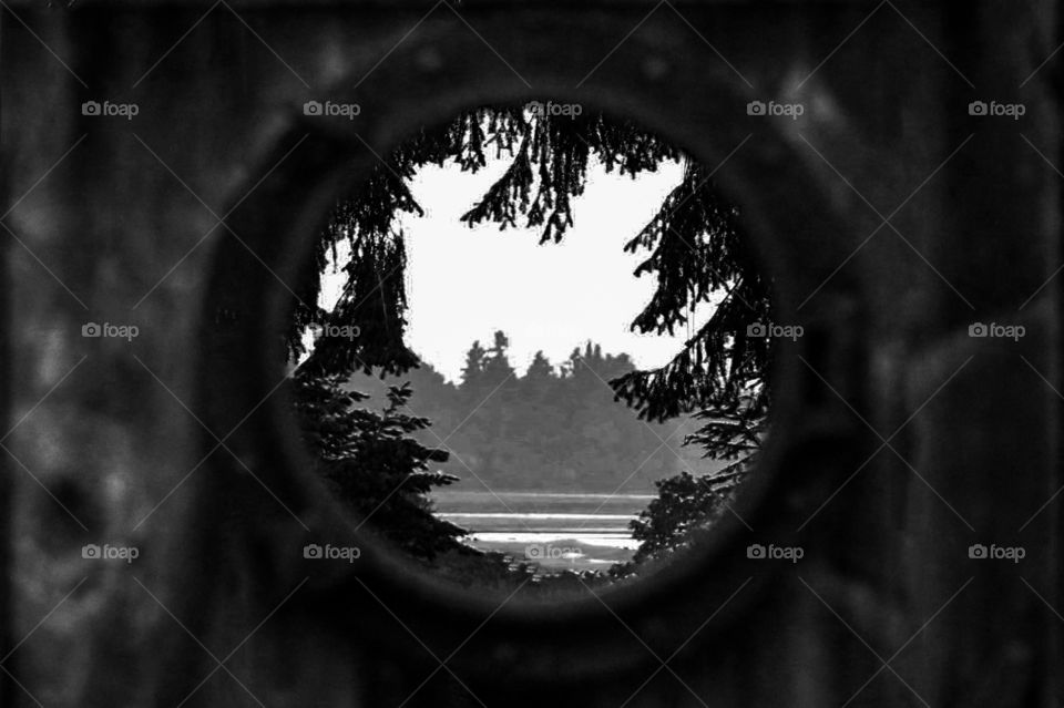 B&W of the view through a ships porthole placed in a wooden yard door. There is a view of the seashore, tidal flats in a small bay, tall trees in the background & the nearby yard tree branches frame the picture like a wreath.