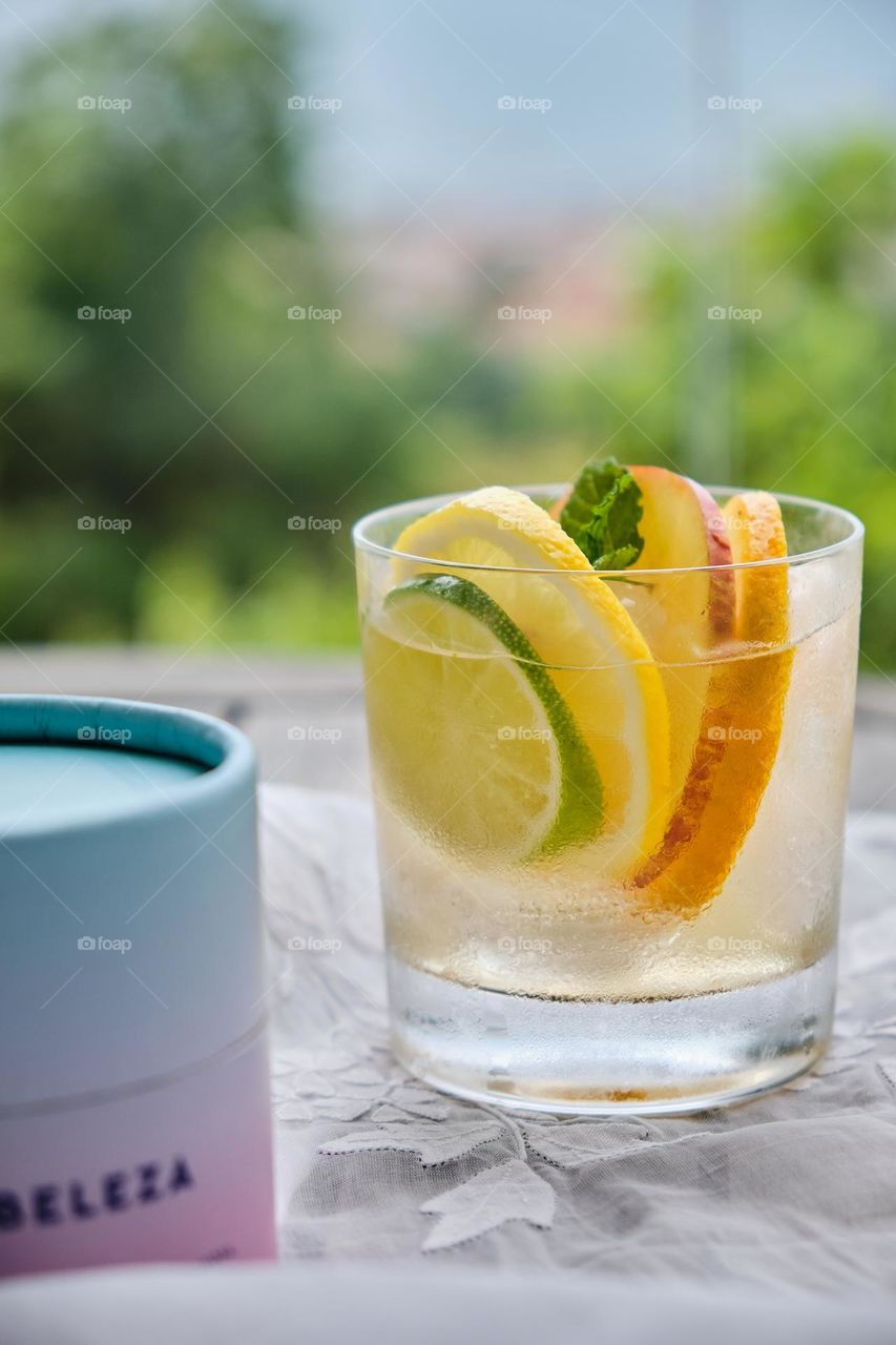 Slices of orange, lime, apple and lemon and a mint leaf inside a transparent cup with water and in foreground pink box with the word beauty