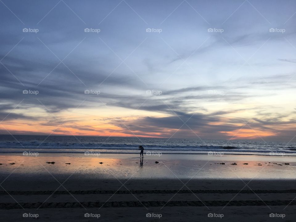 View of the beach, and the sunset. There’s a child standing by the water, and only her silhouette can be seen.